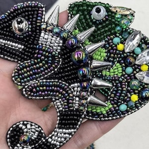 May include: Close-up of two beaded chameleon brooches. One is black with iridescent beads, silver spikes, and a silver eye. The other is green with turquoise, yellow, and clear gem accents, and silver spikes. The brooches are handmade.