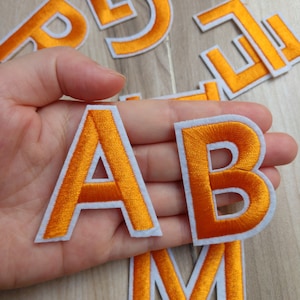 May include: Orange embroidered fabric letters with white borders. The letters A and B are held in a hand, with other letters scattered in the background. The letters are a bright orange colour.