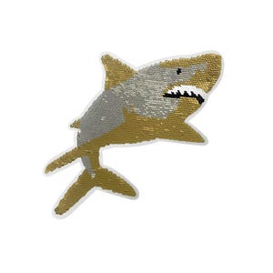 May include: A shark-shaped iron-on patch covered in reversible sequins. The top half of the shark is silver, while the bottom half, fins, and tail are gold. The shark features a white mouth area with black stitched teeth and a black eye detail.