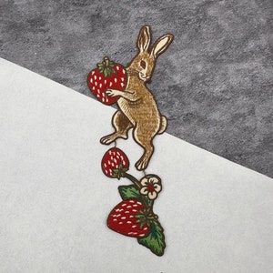 May include: A brown rabbit with a red strawberry in its paw, embroidered on a white fabric. The rabbit is holding the strawberry up to its mouth. The rabbit is surrounded by green leaves and red strawberries.