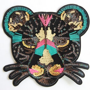 Könnte beinhalten: Ein schwarzer, mit Pailletten besetzter Tierkopf-Patch mit türkisfarbenen, rosafarbenen und goldenen Akzenten. Der Patch ist so gestaltet, dass er wie ein Panther oder Leopard aussieht.