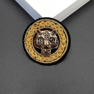 Puede incluir: Un parche bordado en oro con la cabeza de un tigre sobre un fondo de terciopelo negro. La cabeza del tigre está rodeada por un diseño circular de hilo dorado.