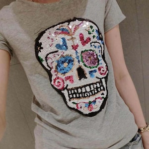 Könnte beinhalten: Graues T-Shirt mit einem bunten Pailletten-Schädel-Design. Der Schädel hat weiße, blaue, rosa und grüne Akzente mit schwarzer Kontur. Das Shirt hat kurze Ärmel und einen Rundhalsausschnitt.