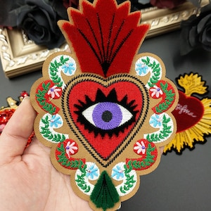 May include: Embroidered patch with a red heart and purple eye, encircled by floral and leaf patterns. The patch has a tan border and a red flame design at the top. The patch is held in a hand.