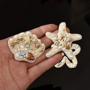 May include: Two decorative seashell and starfish embellishments. The seashell is shaped like a fan and the starfish has five points. Both are adorned with pearls, small shells, and rhinestones. The embellishments are on a white base with a silver border.