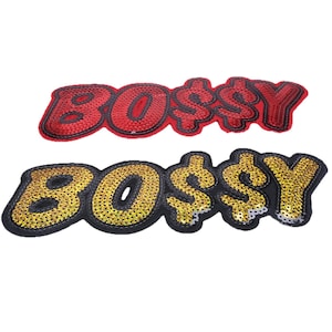 Puede incluir: Dos parches bordados con la palabra BOSSY, donde el signo de dólar reemplaza la letra S. Un parche tiene lentejuelas rojas sobre fondo rojo y el otro tiene lentejuelas doradas sobre fondo negro. Ambos parches tienen la forma del texto.
