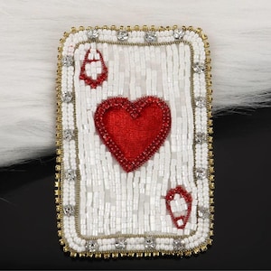 Puede incluir: Un parche rectangular con cuentas en forma de carta de juego as de corazones. El fondo está hecho de cuentas blancas, con un gran corazón rojo central y símbolos de corazón rojo más pequeños en las esquinas, bordeado por cristales dorados y transparentes.