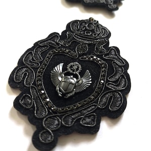 Könnte beinhalten: Zwei schwarze, bestickte Aufnäher mit aufwendigen Designs. Der größere Aufnäher hat eine Schildform mit einem metallenen Skarabäus-Emblem und einer Krone. Die Aufnäher sind mit schwarzen Perlen und detaillierten Nähten verziert.