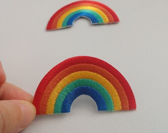 May include: Two embroidered rainbow patches. The patches display a vibrant array of colours, including red, orange, yellow, green, and blue, arranged in a semi-circular shape. The patches are on a light grey background.
