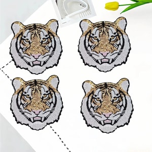 Op de afbeelding: Vier pailletten tijgerkop patches. Elke patch heeft een goudkleurig pailletten gezicht, witte en zwarte details en een roze neus. De patches zijn gerangschikt op een wit oppervlak.