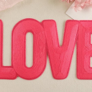 Pode incluir: Letras de feltro rosa "LOVE" com lantejoulas. Cada letra tem aproximadamente 10 cm de altura e 7,5 cm de largura.