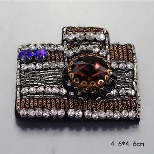 May include: A decorative camera-shaped patch, 4.6 x 4.6 cm, with a black base. The patch is embellished with silver, brown, and black beads, clear and purple rhinestones, and a large, maroon-colored jewel. The camera's dimensions are noted.