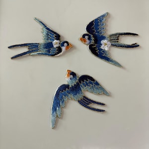 Könnte beinhalten: Drei gestickte Schwalben-Aufnäher in Dunkelblau, Hellblau und Orange. Zwei der Vögel haben ein kleines weißes Blumendetail auf den Flügeln. Jeder Aufnäher ist mit einem metallisch goldenen Faden umrandet, der eine detaillierte Stickerei zeigt.