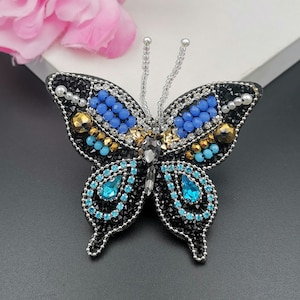 Può includere: Spilla a forma di farfalla nera con intricate perline. Le ali presentano dettagli blu, oro e turchese, con una disposizione centrale di perline trasparenti e argentate. Un accessorio elegante e raffinato per completare qualsiasi look.