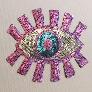 May include: A pink and gold sequin eye patch with a turquoise and black pupil. The eye patch is shaped like a sun with rays coming out from the sides.
