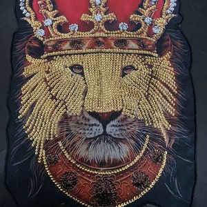 Può includere: Un viso di leone con una corona dorata e una collana dorata, tutto fatto di strass. Il viso del leone è marrone e nero, e lo sfondo è nero.