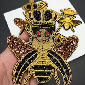 Puede incluir: Parches bordados de abeja con lentejuelas doradas, negras y marrones. Los parches de abeja tienen una corona en la cabeza y ojos rojos. Los parches están sobre una superficie blanca.