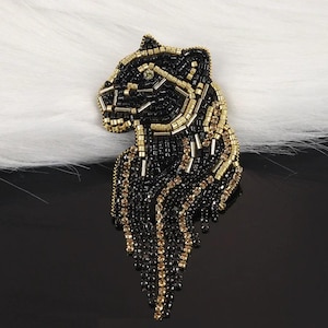 May include: A beaded brooch in the shape of a panther's head. The brooch features black and gold beads, with a green gem for the eye. The panther's head is detailed with gold beads outlining the face and mane.