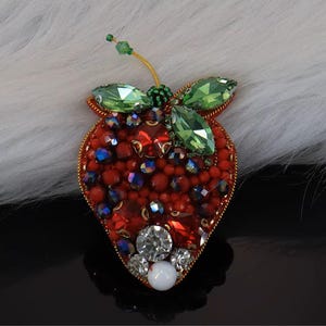 Peut inclure: Broche en forme de fraise avec un contour doré. La broche est recouverte de perles et de pierres précieuses rouges, avec des gemmes vertes en forme de feuilles et une tige en fil d'or. La broche a une perle blanche et des gemmes en cristal transparent.