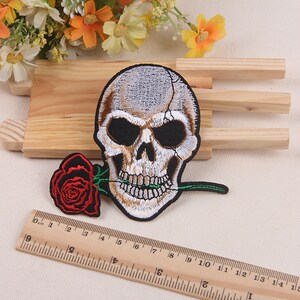 May include: Embroidered iron-on patch featuring a white skull with black eyes and a red rose in its mouth. The skull is biting down on a green stem.