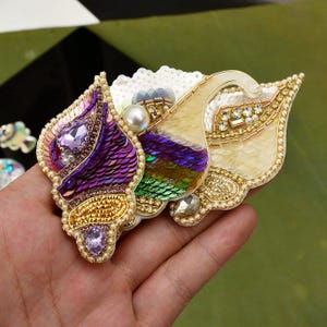 Könnte beinhalten: Eine dekorative Brosche in Form einer Muschel, aufwendig verziert mit violetten, grünen und cremefarbenen Pailletten, Goldperlen, einer großen weißen Perle sowie facettierten violetten und klaren Kristallen in einem detaillierten Muster.