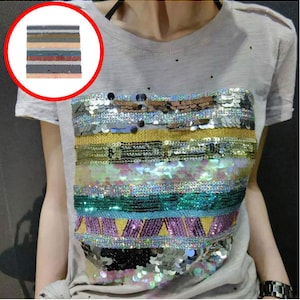 Puede incluir: Una camiseta blanca con un diseño de lentejuelas de colores en la parte delantera. Las lentejuelas están dispuestas en rayas horizontales y un patrón geométrico.