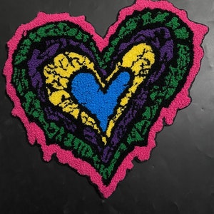 以下が含まれることがあります： ピンク、グリーン、パープル、イエロー、ブルーの糸で編まれた、カラフルなハート型のラグ。ラグは、重ねられたデザインになっています。