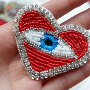 May include: A heart-shaped appliqué with a beaded design. It features a blue eye, red detailing, and a border of silver rhinestones. Suitable for clothing or accessories.