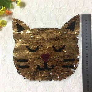 May include: A gold sequined cat face patch with black eyes, whiskers, and a nose. The patch is approximately 13 cm wide and 13 cm tall. The cat's face is centered on a white background.
