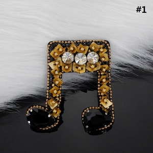 Puede incluir: Un broche de nota musical dorada y negra con diamantes de imitación. El broche está decorado con lentejuelas doradas y cuentas negras.