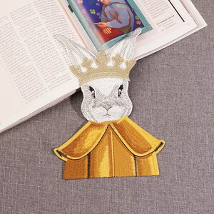 May include: A white embroidered rabbit wearing a gold crown and a yellow cape. The rabbit has a serious expression on its face.