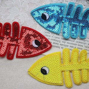 Può includere: Tre toppe termoadesive a forma di pesce con squame di paillettes. I pesci sono rosso, blu e giallo. Ogni pesce ha un design a scheletro e un occhio grande.