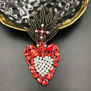 May include: A red and white heart-shaped rhinestone appliqué with a gold cross and flame design. The appliqué is ideal for adding a touch of sparkle to clothing, bags, or other crafts.