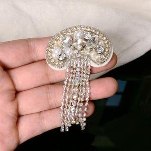 May include: A semicircular brooch featuring a white base adorned with clear faceted crystals, white faux pearls, and a silver-toned border. Multiple strands of shimmering, iridescent crystal fringe hang from the bottom of the brooch.