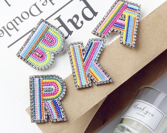 Peut inclure: Quatre lettres brodées colorées, B, R, A et K, avec des strass, sont disposées sur un sac en papier brun. Les lettres sont faites de fil de couleur arc-en-ciel et ont un contour blanc. Les lettres mesurent environ 5 cm de haut.