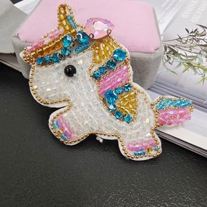 Op de afbeelding: Een witte unicorn applicatie met roze, blauwe en gouden kralen. De unicorn heeft een grote, regenboogkleurige roze edelsteen op zijn voorhoofd.