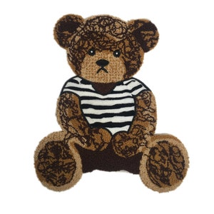 May include: A brown teddy bear rug with a black and white striped shirt. The bear has a light brown face and paws, with darker brown detailing on the body and ears. The rug is made of a soft, textured material.