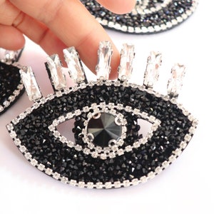 May include: Black and silver eye-shaped rhinestone embellishment with a large black rhinestone in the center. The embellishment is surrounded by smaller rhinestones.