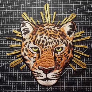 May include: Embroidered iron-on patch featuring a detailed illustration of a leopard's face with a golden sunburst behind it.