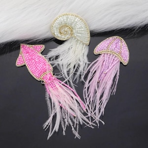 Puede incluir: Tres adornos de criaturas marinas con cuentas y plumas. Un calamar rosa, una medusa morada y una concha blanca.