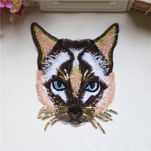 Puede incluir: Primer plano de un rostro de gato marrón y blanco bordado con lentejuelas. Los ojos del gato son azules y el pelaje está detallado con hilo dorado.