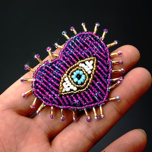 May include: A purple heart-shaped beaded patch with a gold-rimmed blue evil eye in the centre. The patch has gold-coloured spikes around the edges.
