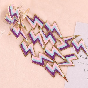 May include: A collection of colourful lightning bolt patches. Each patch features a zig-zag design with purple, light blue, and pink stripes, outlined with gold glitter. The patches are arranged on a light pink surface.