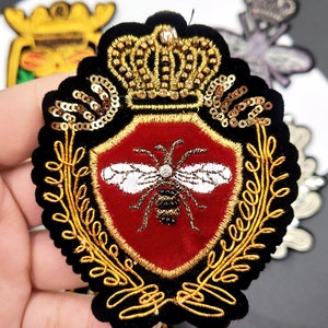 Puede incluir: Un parche de terciopelo negro con un escudo de terciopelo rojo que presenta una abeja bordada en oro. El escudo está rodeado de hojas bordadas en oro y una corona dorada con diamantes de imitación.
