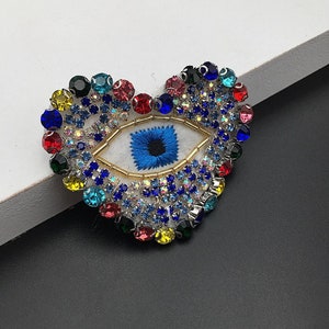 May include: A heart-shaped patch with a blue evil eye embroidered in the centre. The heart is covered in colourful rhinestones.