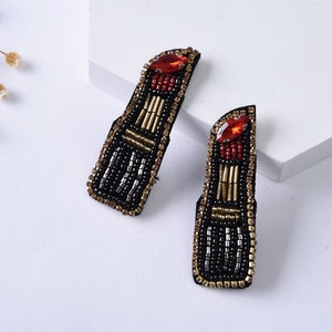 May include: A pair of black beaded earrings shaped like lipstick tubes. Each earring features a red rhinestone and gold beads.