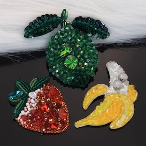 May include: Three beaded fruit brooches: a green lime, a red strawberry, and a yellow banana. The lime has green leaves and a large green gem. The strawberry has a green stem and red gems. The banana is peeled back, revealing white and silver details.