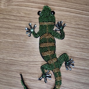 May include: A beaded lizard applique in shades of green and gold, with black accents. The lizard features intricate beadwork, including small black beads for eyes and sparkling embellishments on its feet. The applique is set against a wooden surface.