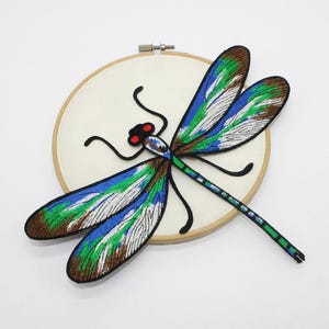 May include: A colourful embroidered dragonfly, featuring brown, green, blue, and white wings, black antennae, and red eyes. The dragonfly is stitched onto cream fabric, held within a wooden embroidery hoop. The design is detailed.