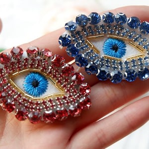May include: Two embroidered evil eye charms with blue and red rhinestones. The eyes are white with blue pupils and gold accents.